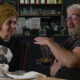 Two people sit at a cafe table and chat; a woman with a mustard head wrap smiles while a bearded man gestures with his hands.