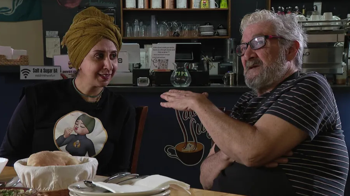 Two people sit at a cafe table and chat; a woman with a mustard head wrap smiles while a bearded man gestures with his hands.