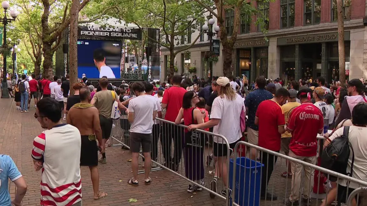 Crowd gathered behind metal barricades watching a large outdoor screen at a street event in Pioneer Square.