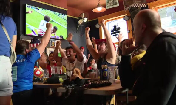 Group of fans at a bar cheering with arms raised as a soccer match plays on a TV screen above them.