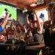 Group of fans at a bar cheering with arms raised as a soccer match plays on a TV screen above them.