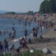 Crowded sandy beach along a calm shore with people swimming, wading, and sunbathing under umbrellas and trees in the distance.