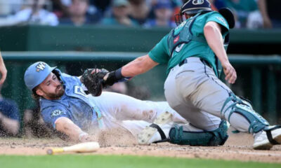 Baseball play: a blue-uniformed runner slides headfirst toward a tag as the teal-caught catcher reaches with his glove, dust rising and a bat on the dirt nearby.