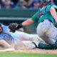 Baseball play: a blue-uniformed runner slides headfirst toward a tag as the teal-caught catcher reaches with his glove, dust rising and a bat on the dirt nearby.