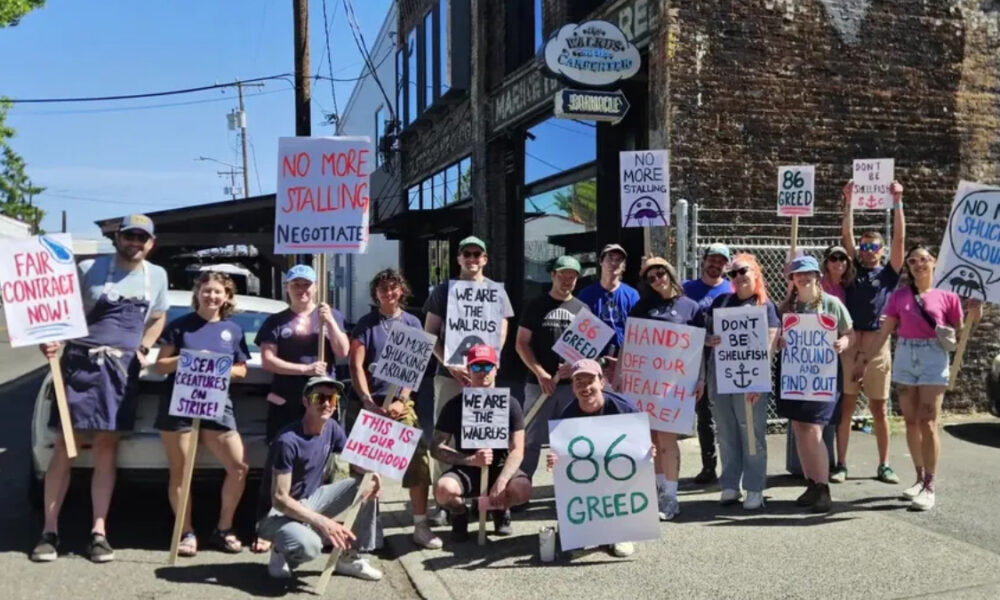 Group of protesters on a sunny street holding handmade signs advocating fair contracts and opposing greed and healthcare cuts.