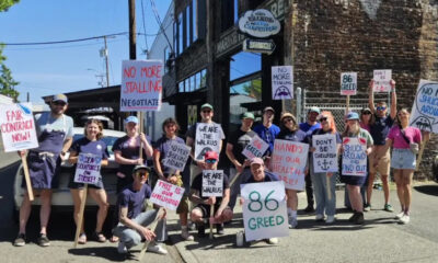 Group of protesters on a sunny street holding handmade signs advocating fair contracts and opposing greed and healthcare cuts.