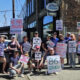 Group of protesters on a sunny street holding handmade signs advocating fair contracts and opposing greed and healthcare cuts.