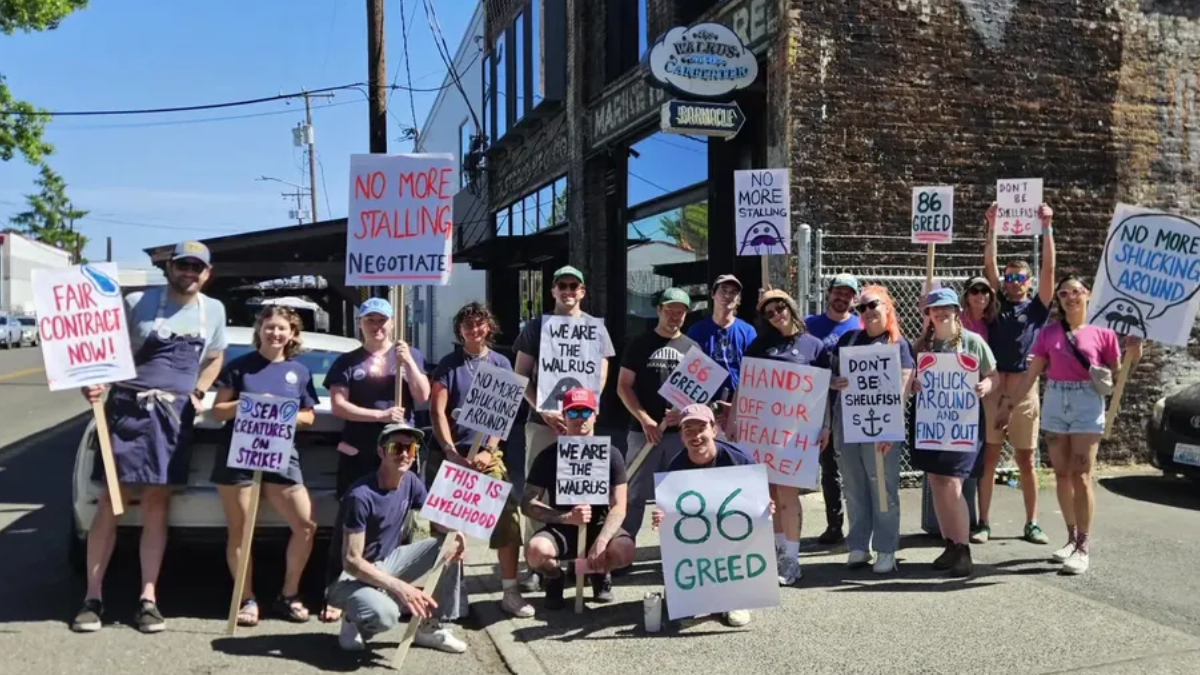 Group of protesters on a sunny street holding handmade signs advocating fair contracts and opposing greed and healthcare cuts.
