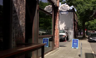 Urban sidewalk scene with brick storefronts and a blue sidewalk sign in front of a cafe/bar named 'Barnacle'.