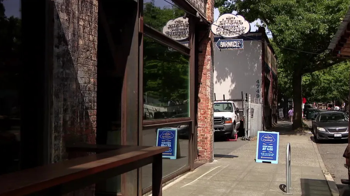 Urban sidewalk scene with brick storefronts and a blue sidewalk sign in front of a cafe/bar named 'Barnacle'.