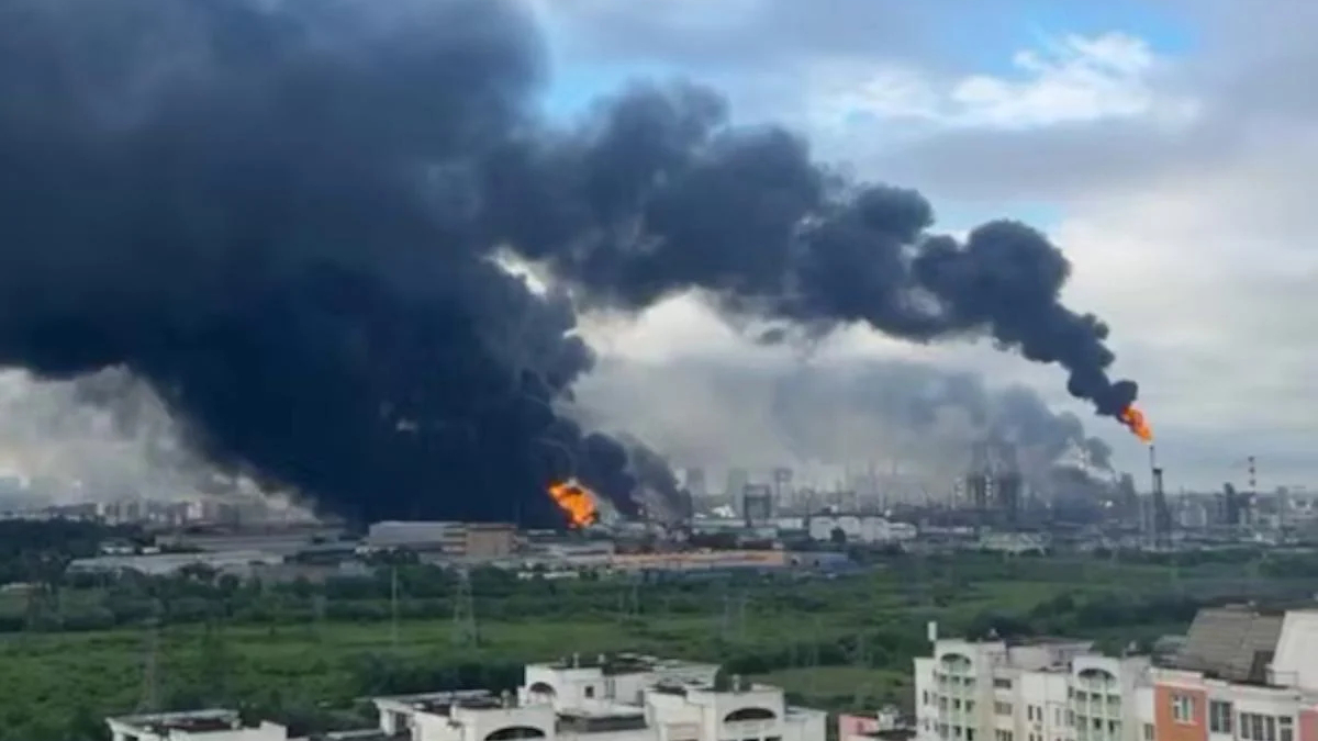 Large plume of dark smoke rising from an industrial complex with flames, near a cityscape.