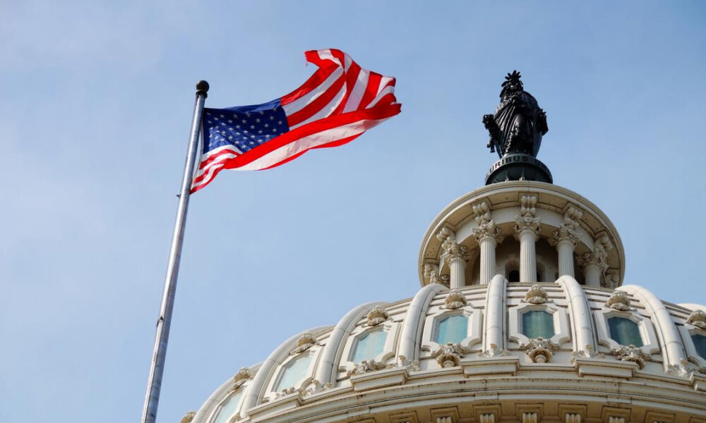 American flag waving on a flagpole beside an ornate domed government building with a statue atop it in a blue sky.
