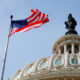 American flag waving on a flagpole beside an ornate domed government building with a statue atop it in a blue sky.