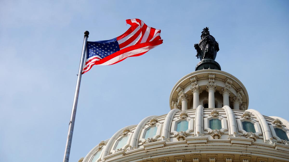 American flag waving on a flagpole beside an ornate domed government building with a statue atop it in a blue sky.