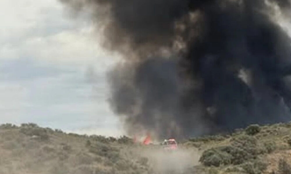 A brush fire on a hillside with a large black smoke plume and a red emergency vehicle nearby