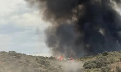 A brush fire on a hillside with a large black smoke plume and a red emergency vehicle nearby