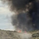 A brush fire on a hillside with a large black smoke plume and a red emergency vehicle nearby