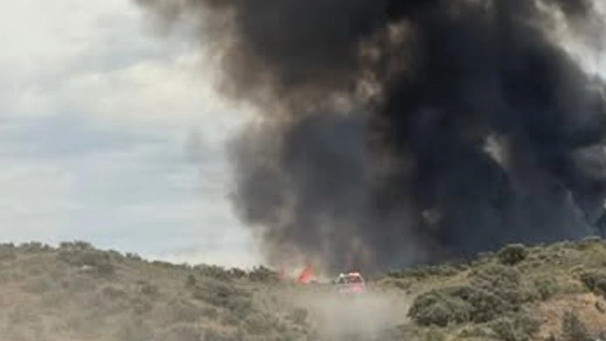 A brush fire on a hillside with a large black smoke plume and a red emergency vehicle nearby