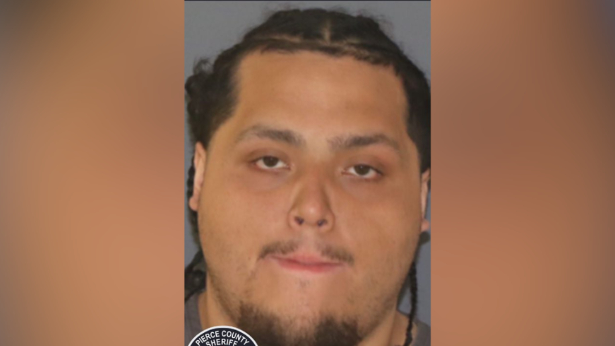 Mugshot of a man with braided hair and goatee against a gray background; a sheriff badge is partially visible in the corner.