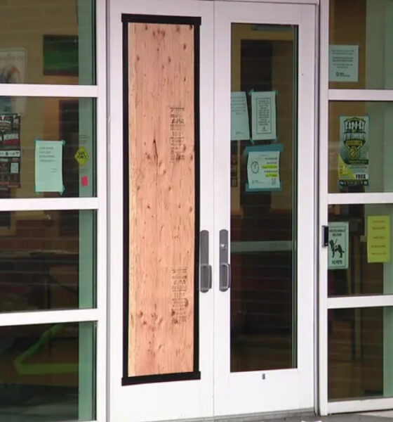 Glass double doors with a plywood panel taped over the center; entry blocked, with notices on the glass and a brick exterior wall nearby, plus a bright green scooter to the right.