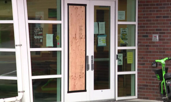 Glass double doors with a plywood panel taped over the center; entry blocked, with notices on the glass and a brick exterior wall nearby, plus a bright green scooter to the right.