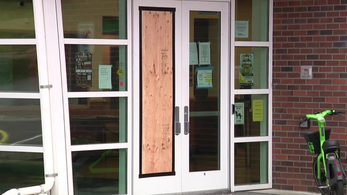 Glass double doors with a plywood panel taped over the center; entry blocked, with notices on the glass and a brick exterior wall nearby, plus a bright green scooter to the right.