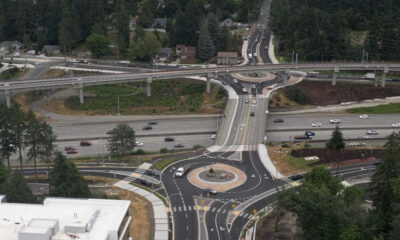 Aerial view of a multi-level highway interchange with a roundabout at the center, surrounding roads with moving cars and trees in a suburban area.