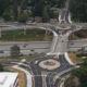 Aerial view of a multi-level highway interchange with a roundabout at the center, surrounding roads with moving cars and trees in a suburban area.