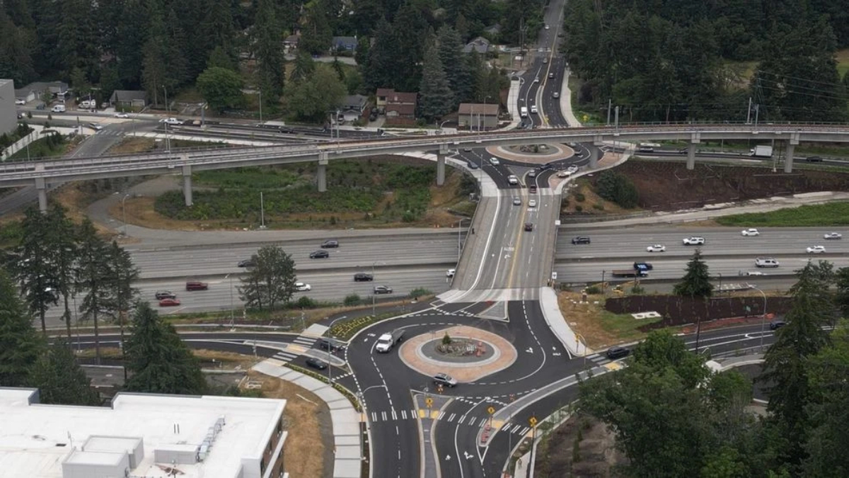 Aerial view of a multi-level highway interchange with a roundabout at the center, surrounding roads with moving cars and trees in a suburban area.