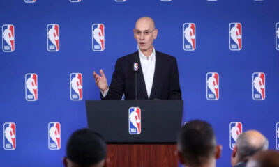 Bald man in a suit speaking at a podium during an NBA press conference, blue backdrop with NBA logos behind him; audience visible in foreground.