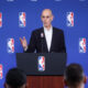 Bald man in a suit speaking at a podium during an NBA press conference, blue backdrop with NBA logos behind him; audience visible in foreground.