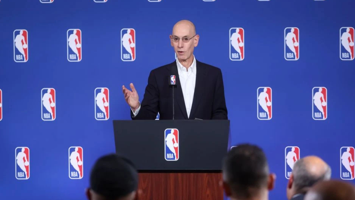 Bald man in a suit speaking at a podium during an NBA press conference, blue backdrop with NBA logos behind him; audience visible in foreground.