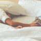 Arm extending from under white sheets to grab a silver alarm clock on a white bed.