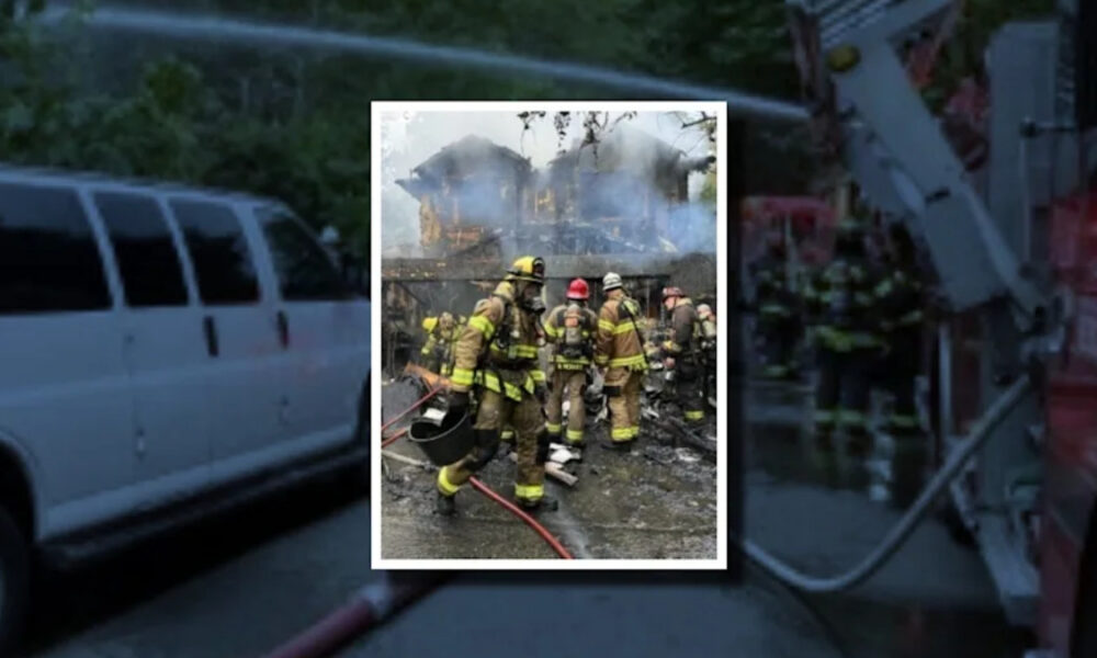 Firefighters in full turnout gear fight a blaze in a burning house with smoke and water on the ground.