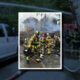 Firefighters in full turnout gear fight a blaze in a burning house with smoke and water on the ground.