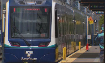 Blue SoundTransit light rail train at a station platform with yellow safety bollards and a waiting passenger in the background.