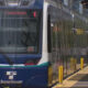 Blue SoundTransit light rail train at a station platform with yellow safety bollards and a waiting passenger in the background.