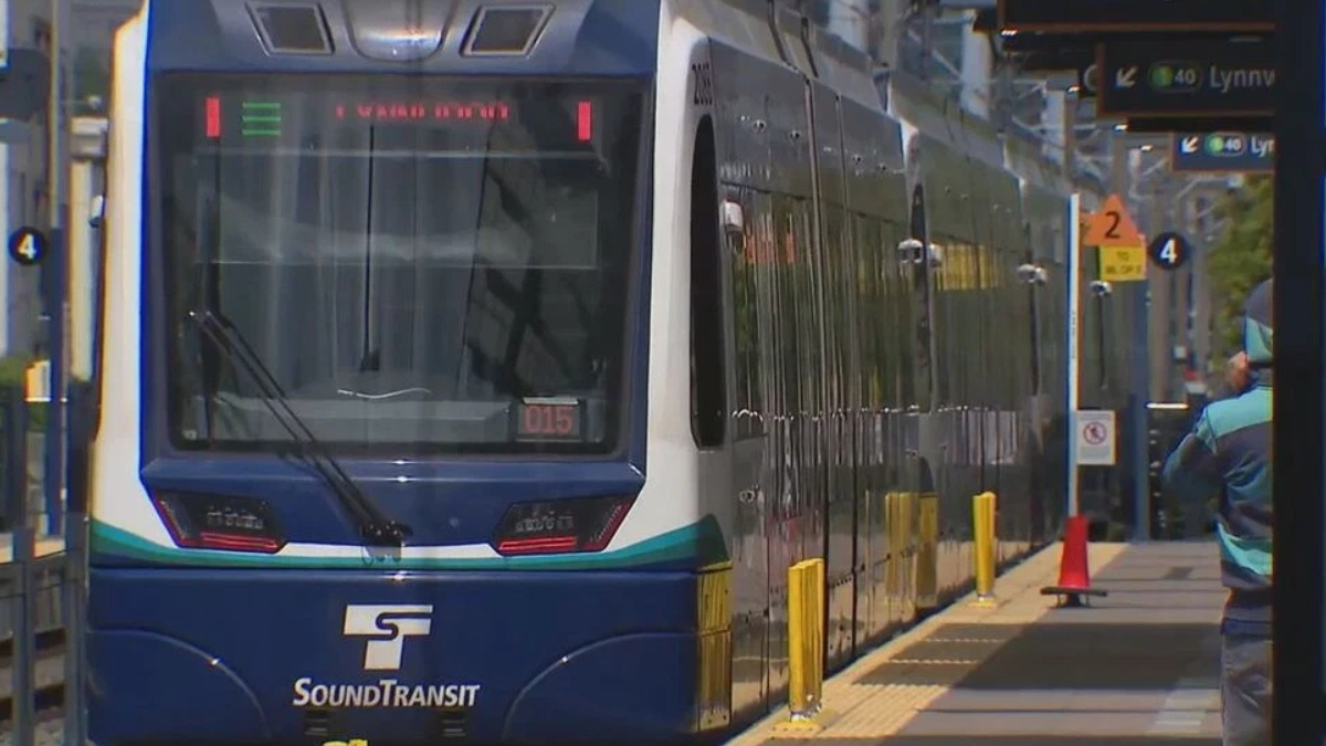 Blue SoundTransit light rail train at a station platform with yellow safety bollards and a waiting passenger in the background.
