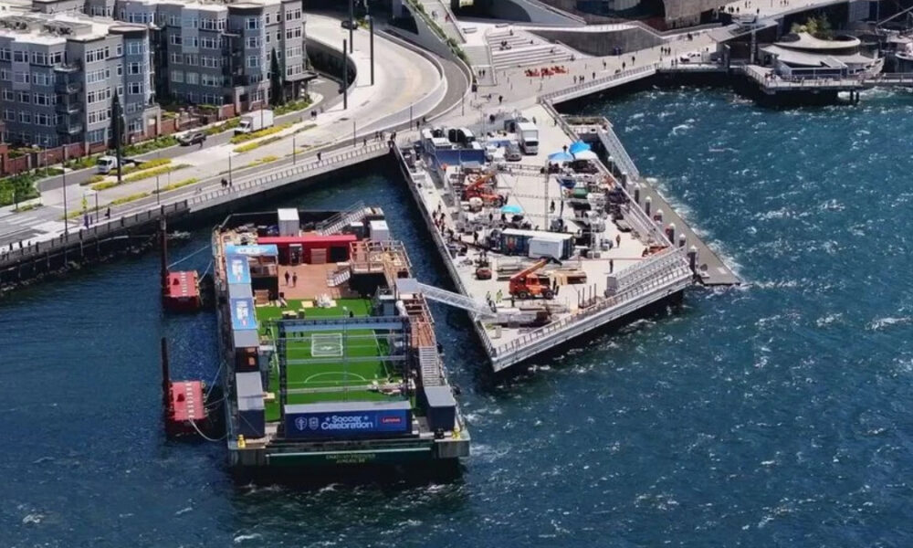Floating soccer field on a barge at a waterfront promenade, with spectators, tents, and equipment on the deck.