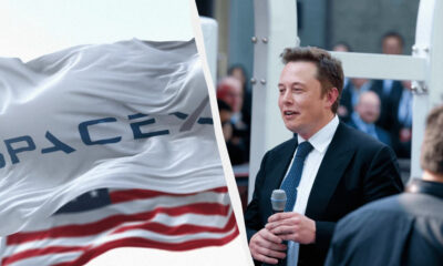 Elon Musk in a suit speaking into a microphone at a SpaceX event, with a SpaceX flag in the foreground.