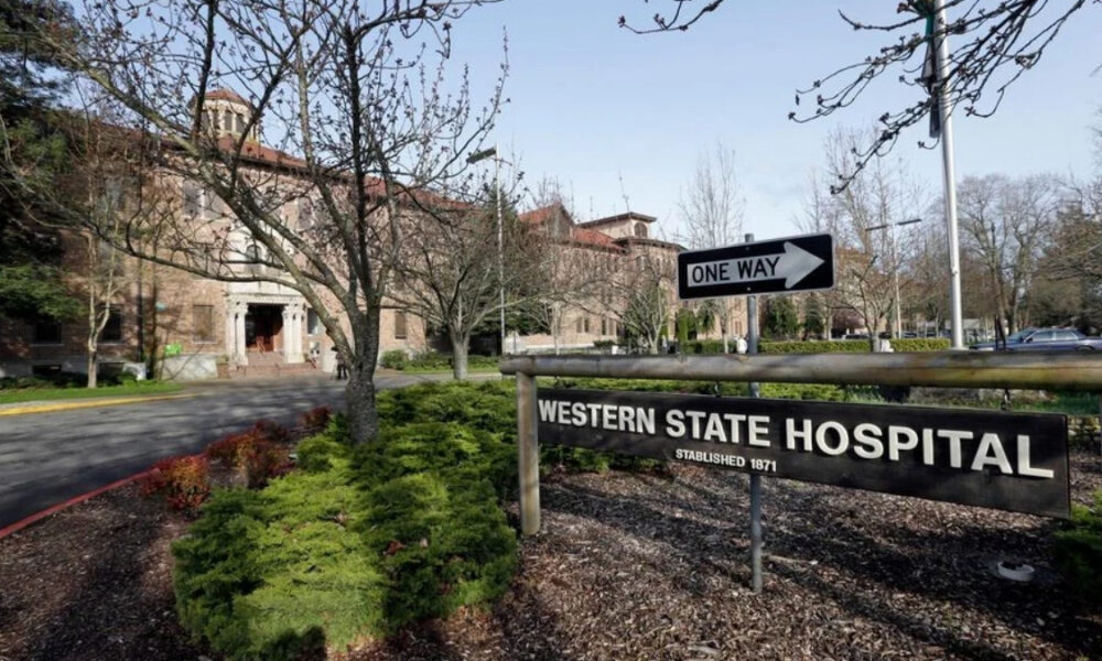Western State Hospital sign in the foreground with a one-way arrow sign and a brick campus building behind, surrounded by trees and shrubs.