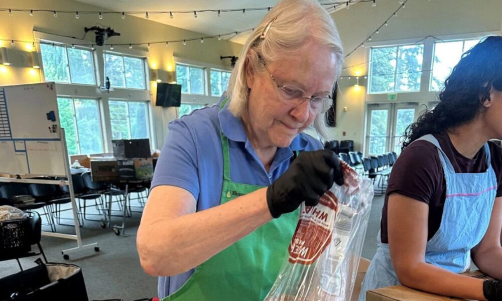Older woman in a blue polo and green apron seals a plastic bag with a red label at a volunteer station in a bright, string-lit room.