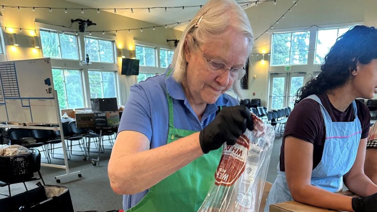 Older woman in a blue polo and green apron seals a plastic bag with a red label at a volunteer station in a bright, string-lit room.