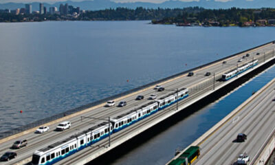 Aerial view of a dual transport corridor: trains on an elevated rail track beside a multi-lane highway over a body of water, with a city in the distance.