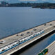 Aerial view of a dual transport corridor: trains on an elevated rail track beside a multi-lane highway over a body of water, with a city in the distance.