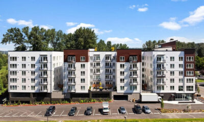 Modern multi-story apartment building with white and brown facade, balconies, and a parking lot in front.