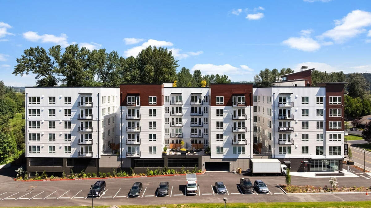 Modern multi-story apartment building with white and brown facade, balconies, and a parking lot in front.