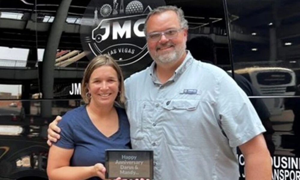 Smiling couple stands in front of a black bus, holding a plaque that reads 'Happy Anniversary Dana & Mandy'.