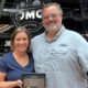 Smiling couple stands in front of a black bus, holding a plaque that reads 'Happy Anniversary Dana & Mandy'.