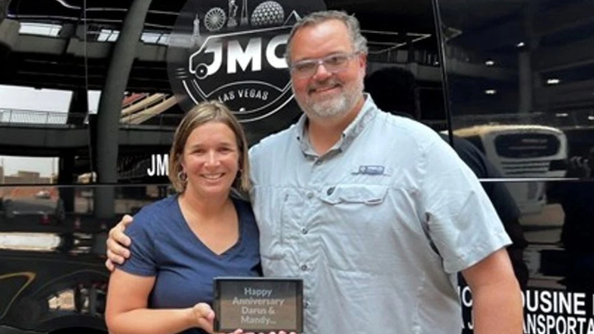 Smiling couple stands in front of a black bus, holding a plaque that reads 'Happy Anniversary Dana & Mandy'.